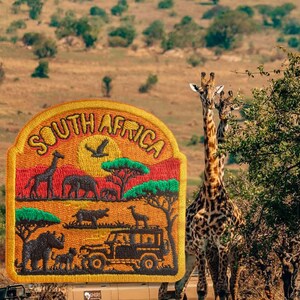 Op de afbeelding: Een halfronde geborduurde patch met de tekst SOUTH AFRICA in gele letters. Het ontwerp toont een savannelandschap met een giraf, olifant, neushoorn en safarivoertuig in tinten oranje, rood, groen en bruin.
