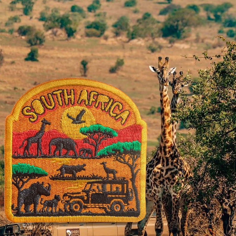 Op de afbeelding: Een halfronde geborduurde patch met de tekst SOUTH AFRICA in gele letters. Het ontwerp toont een savannelandschap met een giraf, olifant, neushoorn en safarivoertuig in tinten oranje, rood, groen en bruin.