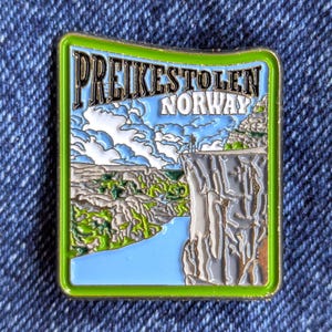 May include: Enamel pin depicting Preikestolen, Norway. The pin has a green border and shows a cliff edge with a person, overlooking a valley and water. The text "PREIKESTOLEN NORWAY" is at the top. The design is in shades of blue, green, and grey.