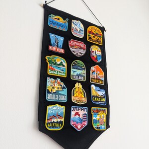 May include: A black fabric wall hanging displaying colorful embroidered travel patches. Patches include destinations like New York, Japan, and Austria. The patches are in various shapes and sizes, with detailed illustrations and text.