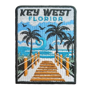 以下が含まれることがあります： キーウェスト、フロリダのシーンを描いた刺繍パッチ。ヤシの木、遊歩道、海の波、そして白と青で「KEY WEST FLORIDA」の文字がデザインされています。パッチには黒い縁取りがあります。