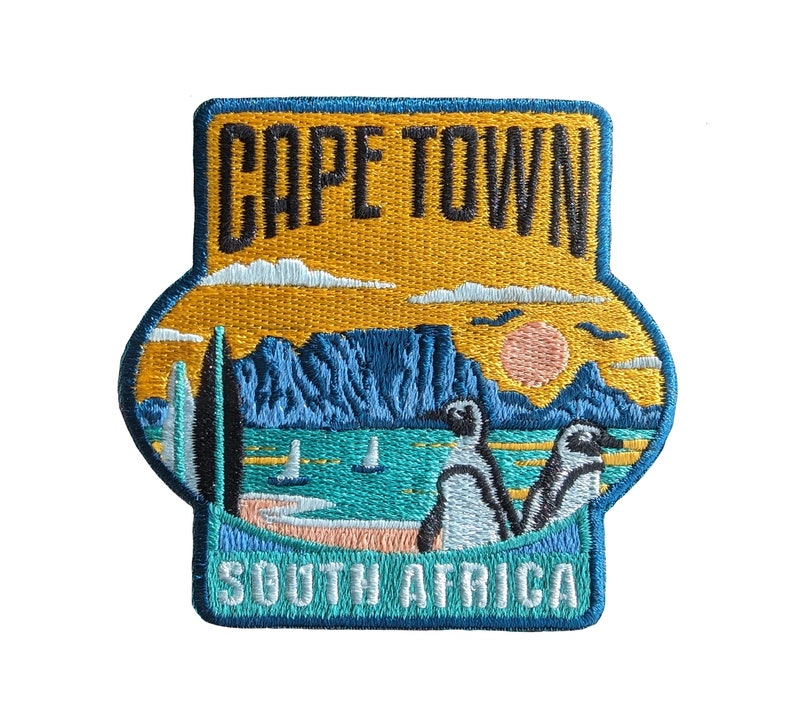 Może przedstawiać: Naszywka haftowana w kolorze niebieskim, ż&oacute;łtym i białym. Zawiera napis "CAPE TOWN" i "SOUTH AFRICA" wraz z pasmem g&oacute;rskim, zachodem słońca i dwoma pingwinami.