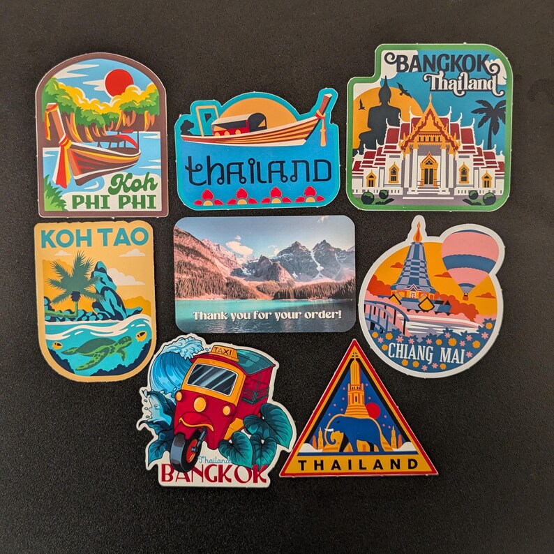 May include: A set of colorful stickers featuring various scenes from Thailand. The stickers include images of temples, boats, tuk-tuks, and landscapes. The text on the stickers includes "Koh Phi Phi", "Koh Tao", "Thailand", "Bangkok", and "Chiang Mai".