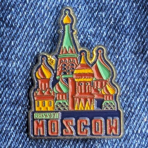 Può includere: Spilla smaltata colorata raffigurante la Cattedrale di San Basilio a Mosca. La spilla presenta dettagli rossi, gialli, verdi e blu, con le parole "MOSCA" e "RUSSIA" su uno stendardo rettangolare.