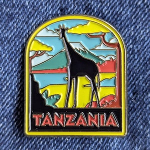 Puede incluir: Un colorido pin de esmalte con la silueta de una jirafa sobre un paisaje con una montaña, árboles y nubes. La palabra "TANZANIA" se muestra en letras rojas en la parte inferior del pin, sobre un fondo negro.