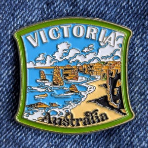 May include: A colorful enamel pin featuring a coastal scene with the text "Victoria Australia." The pin has a green border, a blue sky with white clouds, and a beach with rock formations. The design is detailed and vibrant.