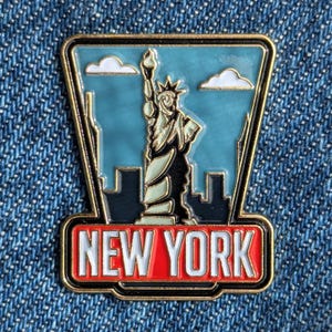 Peut inclure: Une épingle émaillée de New York représentant la Statue de la Liberté sur fond de ciel bleu avec des nuages blancs. L'épingle a une bordure dorée et les mots "NEW YORK" dans un rectangle rouge.