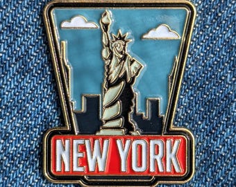 May include: A New York City enamel pin featuring the Statue of Liberty against a blue sky with white clouds. The pin has a gold border and the words "NEW YORK" in a red rectangle.