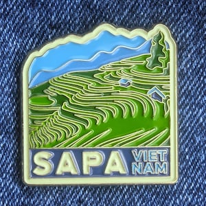 May include: A rectangular enamel pin featuring a landscape of green terraced fields, blue sky, and small houses. The pin has the text "SAPA VIET NAM" in a light yellow font with a dark blue background and a light yellow border.