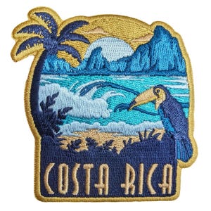 Puede incluir: Un parche bordado que muestra una escena de playa tropical con una palmera, olas del mar, montañas y un tucán. El diseño incluye un sol azul claro y el texto COSTA RICA en letras color canela en la parte inferior, bordeado con hilo dorado.