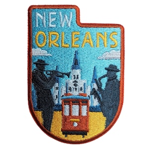 May include: An embroidered patch featuring the text NEW ORLEANS in yellow and white. The design depicts two dark blue figures playing a trumpet and saxophone, a red streetcar, and a white building with spires against a light blue sky.