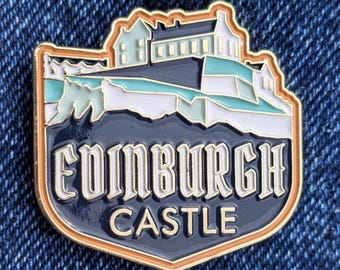 May include: A colorful enamel pin featuring Edinburgh Castle. The pin has a gold border and a navy blue background with the words "Edinburgh Castle" in white. The castle illustration includes white, teal, and light brown accents.
