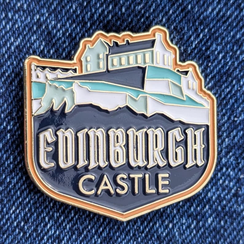 May include: An enamel pin depicting Edinburgh Castle. The pin has a gold-coloured border and a navy blue background. The words "Edinburgh Castle" are in white. The castle illustration includes white, teal, and light brown accents.