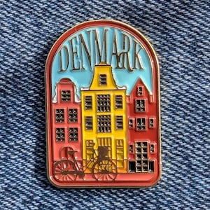 Puede incluir: Un colorido pin de esmalte con una representación estilizada de edificios en Dinamarca. El pin tiene un borde dorado e incluye la palabra "DENMARK" en letras doradas sobre un fondo azul claro. Los edificios son rojos y amarillos, con una bicicleta en primer plano.