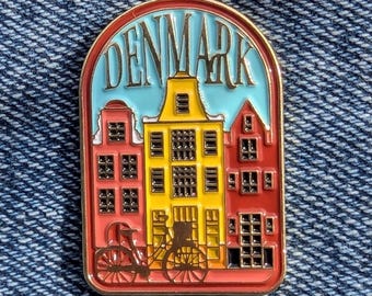 May include: A colorful enamel pin featuring a stylized depiction of buildings in Denmark. The pin has a gold border and includes the word "DENMARK" in gold lettering against a light blue background. The buildings are red and yellow, with a bicycle in the foreground.