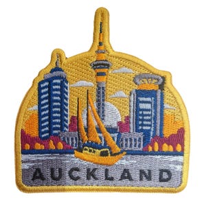 May include: An embroidered patch featuring the Auckland skyline with the Sky Tower, blue skyscrapers, and a yellow sailboat on water. The design includes a yellow border and the word AUCKLAND in black capital letters at the bottom of the patch.