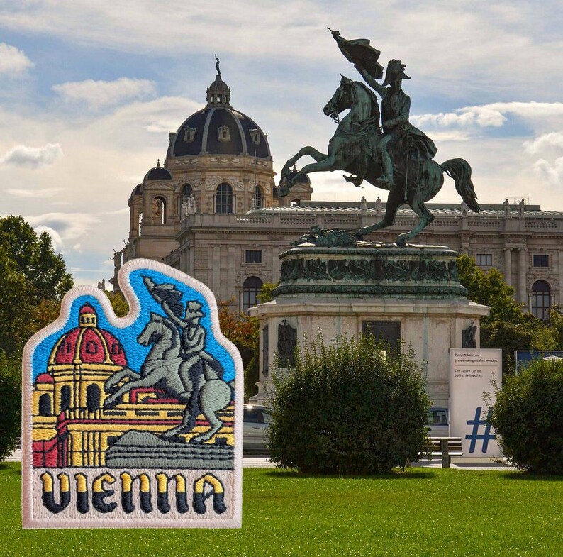 Może przedstawiać: Naszywka haftowana w kolorze białym i beżowym z kolorową ilustracją budynku i powozu ciągniętego przez konie. Tekst "Vienna" jest wyszyty czarnymi, ż&oacute;łtymi i czerwonymi literami na naszywce.