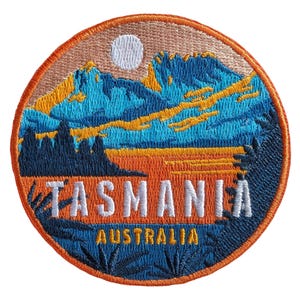 Könnte beinhalten: Ein runder gestickter Aufnäher mit einer Landschaft aus blauen Bergen, einer weißen Sonne und orangefarbenen Feldern. Der Text TASMANIA ist in weißen Großbuchstaben in der Mitte eingestickt, darunter das Wort AUSTRALIA in Gelb, alles orange umrandet.