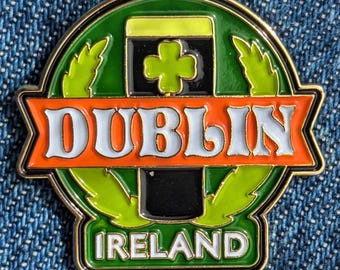 May include: A round, enamel pin with a gold border. The pin features the word "DUBLIN" in white letters on an orange banner. Below the banner is "IRELAND" in white letters. The pin has a green background with a black and gold beer glass and a shamrock.