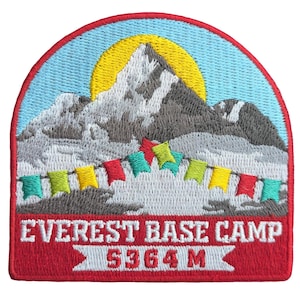 Puede incluir: Parche bordado que representa una cordillera con un sol amarillo y una guirnalda de banderas de colores. El texto "EVEREST BASE CAMP 5364 M" se muestra en una pancarta roja. El parche tiene un fondo de cielo azul y un borde rojo.