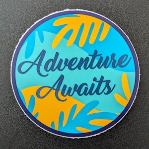May include: Circular sticker with a dark blue border. The text "Adventure Awaits" is in a dark blue script. The design includes turquoise and yellow leaf-like shapes.