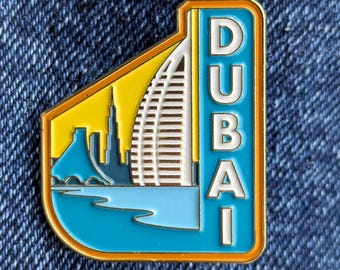 May include: A rectangular enamel pin featuring a Dubai cityscape. The pin has a blue, yellow, and white color scheme with the word "DUBAI" in vertical white letters. The pin includes an image of the Burj Al Arab hotel.