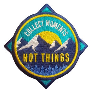 May include: Embroidered patch featuring a mountain and sun design. The patch is navy blue, teal, yellow, and white. Text on the patch reads "COLLECT MOMENTS NOT THINGS". The patch has a compass rose shape.