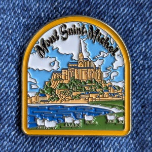 Könnte beinhalten: Bunter Emaille-Pin mit dem Mont Saint Michel in Frankreich. Der Pin hat einen gelben Rand und zeigt die Abtei auf einem grünen Hügel, mit blauem Wasser und Himmel. Der Text "Mont Saint Michel" ist oben gewölbt.