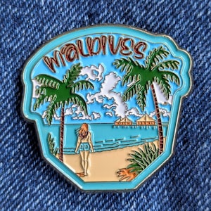 May include: A colorful enamel pin featuring a tropical beach scene with palm trees, a woman, and the word "Maldives" in a stylized font. The pin has a light blue border and a gold-colored metal edge.