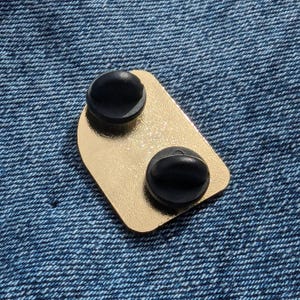 May include: A gold-colored enamel pin with two black pin backs. The pin has a rectangular shape with rounded corners. The pin is set against a blue denim background.