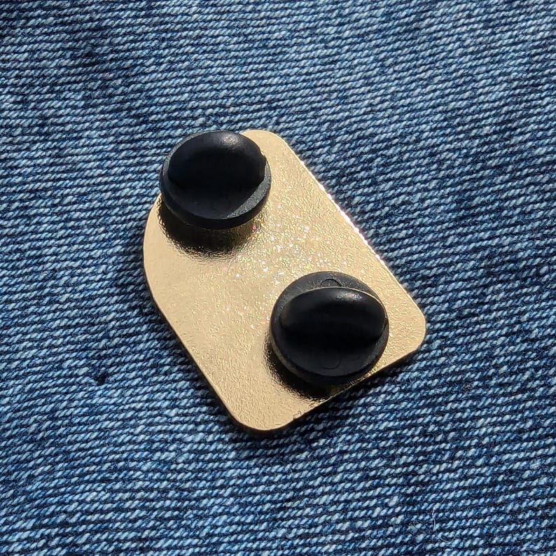 May include: A gold-colored enamel pin with two black pin backs. The pin has a rectangular shape with rounded corners. The pin is set against a blue denim background.