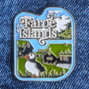 Może przedstawiać: Prostokątna emaliowana przypinka z jasnoniebieską obwódką. Przypinka zawiera napis "Faroe Islands" w białym skrypcie, maskonura i zieloną trawę z czarnymi domami. Niebo jest jasnoniebieskie z białymi chmurami.