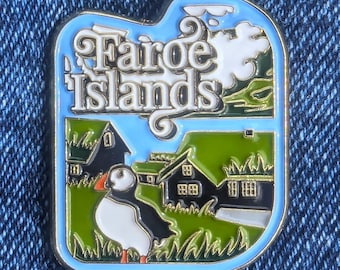 Peut inclure: Épingle en émail rectangulaire avec une bordure bleu clair. L'épingle présente le texte "Faroe Islands" en écriture blanche, un macareux et de l'herbe verte avec des maisons noires. Le ciel est bleu clair avec des nuages blancs.