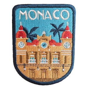 Könnte beinhalten: Gestickter Aufnäher mit dem Wort "MONACO" über einer detaillierten Illustration eines Gebäudes mit zwei Türmen mit roten Spitzen, Palmen und einem blauen Himmelshintergrund. Der Aufnäher hat einen blauen Rand und ein gold-beiges Gebäude.