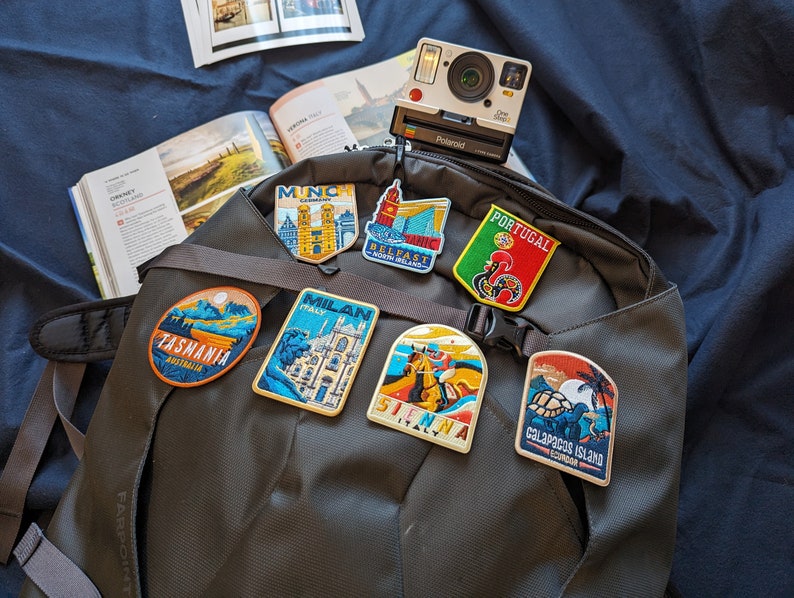 May include: A gray backpack with several colorful embroidered patches attached to it. The patches feature images of various locations, including Tasmania, Milan, Siena, Galapagos Island, Portugal, Munich, and Belfast.