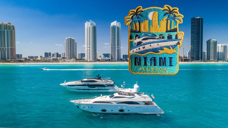 Op de afbeelding: Twee witte jachten liggen aangemeerd in turquoise water voor een skyline. De skyline omvat hoge gebouwen en een strand. Een blauw en geel geborduurd embleem met de tekst "Miami, Florida" bevindt zich in de rechterbovenhoek van de afbeelding.