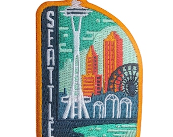 May include: An embroidered patch featuring the Seattle skyline with the Space Needle, orange and red buildings, and a Ferris wheel. The word SEATTLE is stitched vertically in white on a dark blue background, all bordered by a bright orange edge.