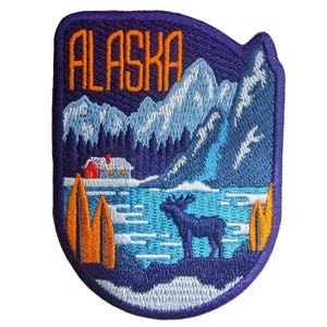 Könnte beinhalten: Gestickter Aufnäher mit einer Alaska-Landschaft. Der Aufnäher hat einen dunkelvioletten Rand und zeigt das Wort "ALASKA" in Orange. Das Design umfasst Berge, einen See, Bäume, einen Elch und eine Hütte mit rotem Dach.