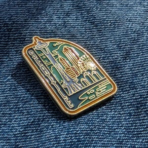 May include: A rectangular enamel pin featuring a Seattle cityscape design. The pin has a gold border and depicts the Space Needle, buildings, and a Ferris wheel in shades of green, blue, and gold. The word "Seattle" is written on the pin.
