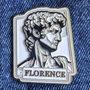 May include: A rectangular enamel pin depicting a profile of Michelangelo's David statue in black, white, and gold. The word "FLORENCE" is displayed in a gold font at the bottom of the pin.