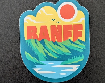 Puede incluir: Una colorida pegatina con la palabra "BANFF" en letras naranjas. La pegatina representa una cordillera, un lago y un sol, todo dentro de un borde azul en forma de nube. Dos pequeños pájaros vuelan en el cielo amarillo.