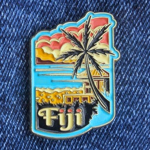 May include: Enamel pin depicting a tropical Fiji scene. The pin has a blue border and shows a palm tree, beach, and sunset. The word "Fiji" is written in a stylized font at the bottom. The pin is set against a denim background.