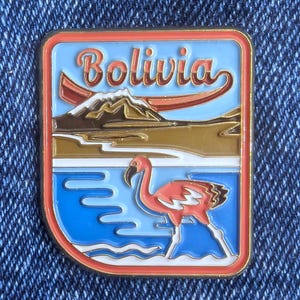 Puede incluir: Un pin rectangular esmaltado con un flamenco en un lago azul, con la palabra "Bolivia" sobre una escena de montaña y barco. El pin tiene un borde rojo y detalles dorados.