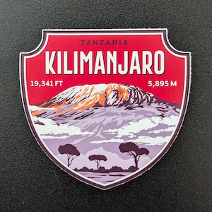May include: A shield-shaped sticker featuring Mount Kilimanjaro in Tanzania. The sticker has a red border with the word "TANZANIA" at the top and "KILIMANJARO" in large white letters. The mountain is depicted with orange and white snow, and the height is listed as 19,341 FT and 5,895 M.