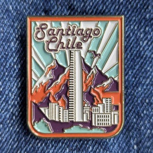 May include: Rectangular enamel pin featuring a colourful design of Santiago, Chile. The pin includes the city name, mountains, a tall building, and a cityscape. The colour palette includes orange, purple, teal, and white.
