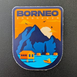 May include: A colorful sticker featuring a Borneo Indonesia landscape. The design includes a mountain range, a setting sun, and small huts on stilts over water. The word "BORNEO" is printed at the top in orange letters.