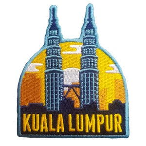 May include: Embroidered patch featuring the Petronas Twin Towers in Kuala Lumpur, Malaysia. The design includes the towers in blue, a yellow and orange sunset, and the city skyline. The text "KUALA LUMPUR" is embroidered in a bold, navy blue font.