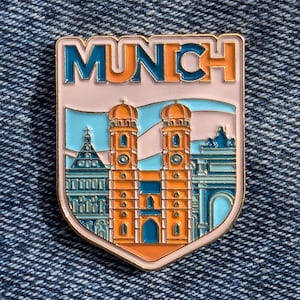 May include: Enamel pin featuring the word "MUNCH" in orange and teal, above a depiction of a city's architecture. The pin has a gold border and is set against a denim background.