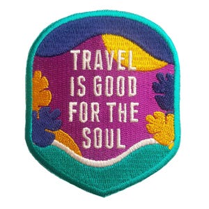 Puede incluir: Parche bordado con el texto "TRAVEL IS GOOD FOR THE SOUL". El parche tiene un fondo morado con detalles amarillos, azules y turquesas. El borde es turquesa y el texto es blanco.
