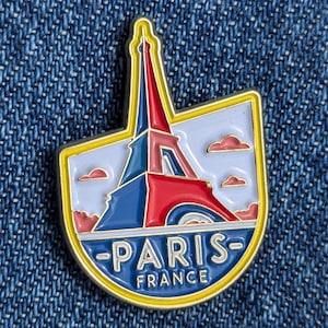 May include: Enamel pin depicting the Eiffel Tower in red, white, and blue, set against a light blue sky with pink clouds. The pin is outlined in gold and has the words "PARIS FRANCE" in gold lettering.